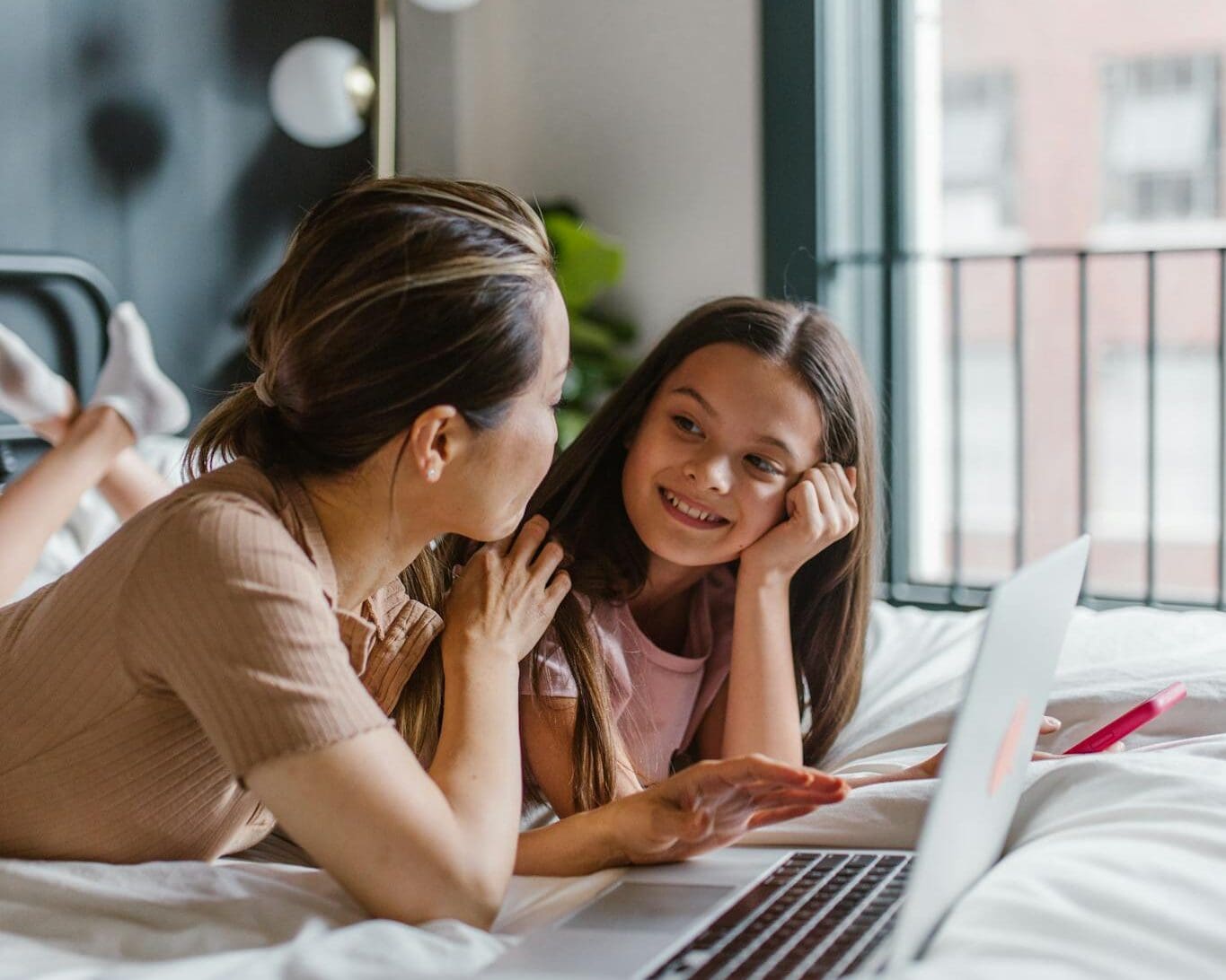 Mother and daughter enjoying quality time using a laptop in a cozy bedroom setting.