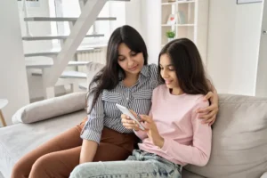 Happy mother and teenage daughter sitting together on couch looking at smartphone, demonstrating positive parent-teen communication about social media safety and digital wellbeing at home